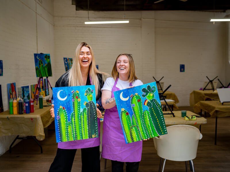 Two women hold up their cactus paintings made at a date night art class.
